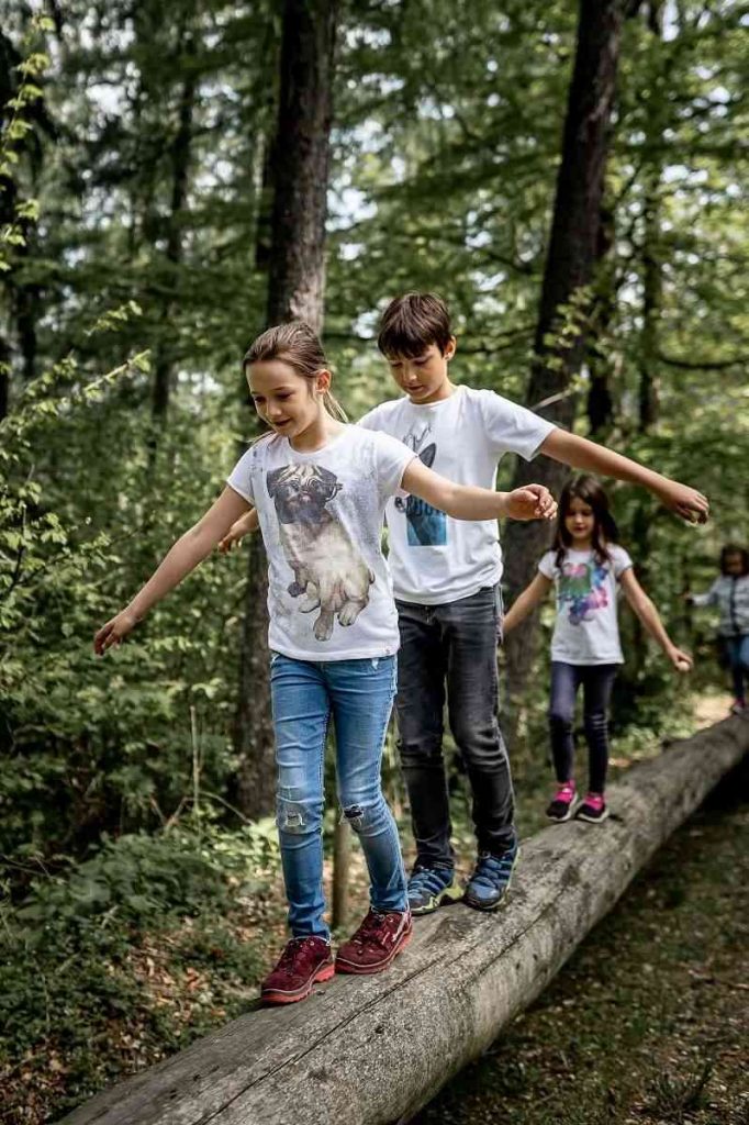 Kinder verschiedenen Alters balancieren auf einem Baumstamm im Zuge des Waldgeburtstags an den sie im Waldpark feiern.