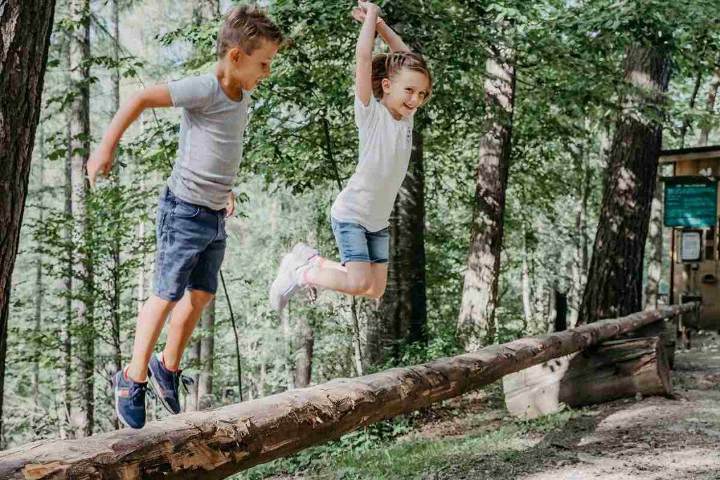 Junge und Mädchen springen über einen Baumstamm beim Ausflug mit ihrer Volksschule