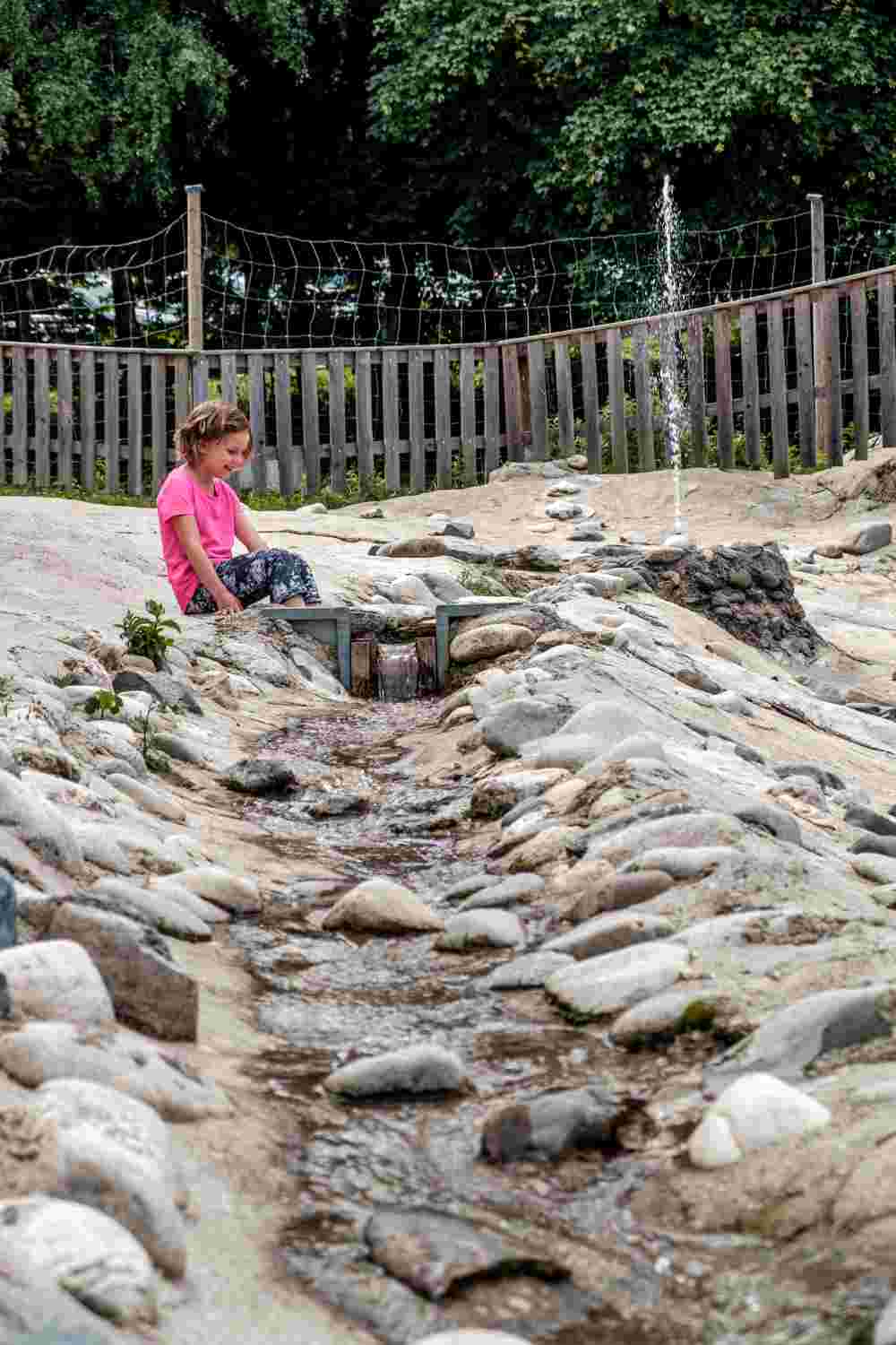 Ein Kind sitzt am Wasserspielplatz auf den Steinen zwischen denen fröhlich das Wasser hinunter plätschert