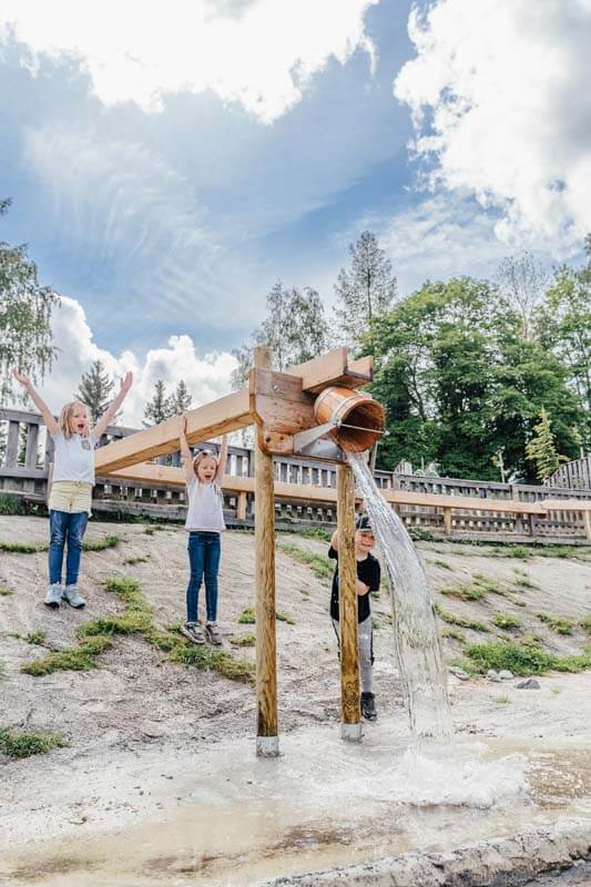 Drei Kinder jubeln beim Entleeren des großen Wasserkübels am Wasserspielplatz in das vorbeiplätschernde Rinnsal