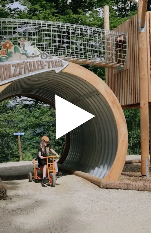 Startbild für Video zum Go-Kart-Fahren im Waldpark. Mädchen kommt mit einem Tretroller durch das Rohr im Eingangsbereich des Areals gefahren.