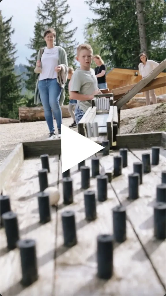 Thumbnail für Video zu Waldkugelbahn auf der Teichalm - am Bild sind Kinder und Erwachsene an einer Station bei der die Kugel durch ein Plateau mit verschiedenen Holzpfeilern rollt und alle schauen zu